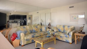 Ein Wohnzimmer mit einer Couch, einem Couchtisch und einem Fernseher. Im Hintergrund ist eine Küchenzeile zu sehen.