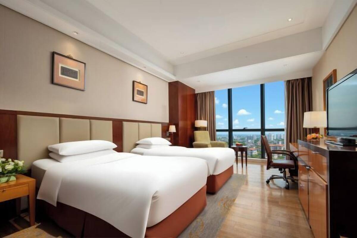 Habitación de hotel con dos camas, un escritorio, una silla, un televisor y un ventanal con vistas a la ciudad.