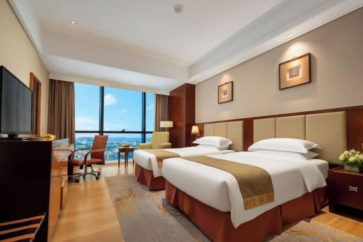 Habitación de hotel con dos camas, un escritorio, una silla, televisión y ventanas amplias con vistas a la ciudad.