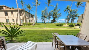 A patio with a table and chairs, overlooking a well-maintained lawn and palm trees, with a building in the background. - Stunning Ocean View Condo  Pono Kai Resort  G106 (Kapa`a)