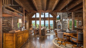 A rustic cabin interior with a stone fireplace, wooden furniture, and large windows offering a mountain view. - Elegant Home with Incredible Views (Banner Elk)