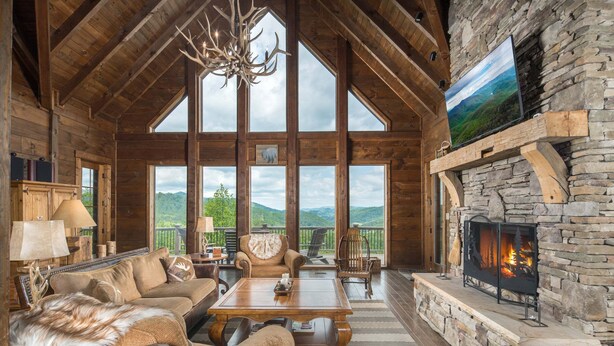 A log cabin interior with a stone fireplace, large windows, a chandelier, and a cozy seating area. - Elegant Home with Incredible Views (Banner Elk)
