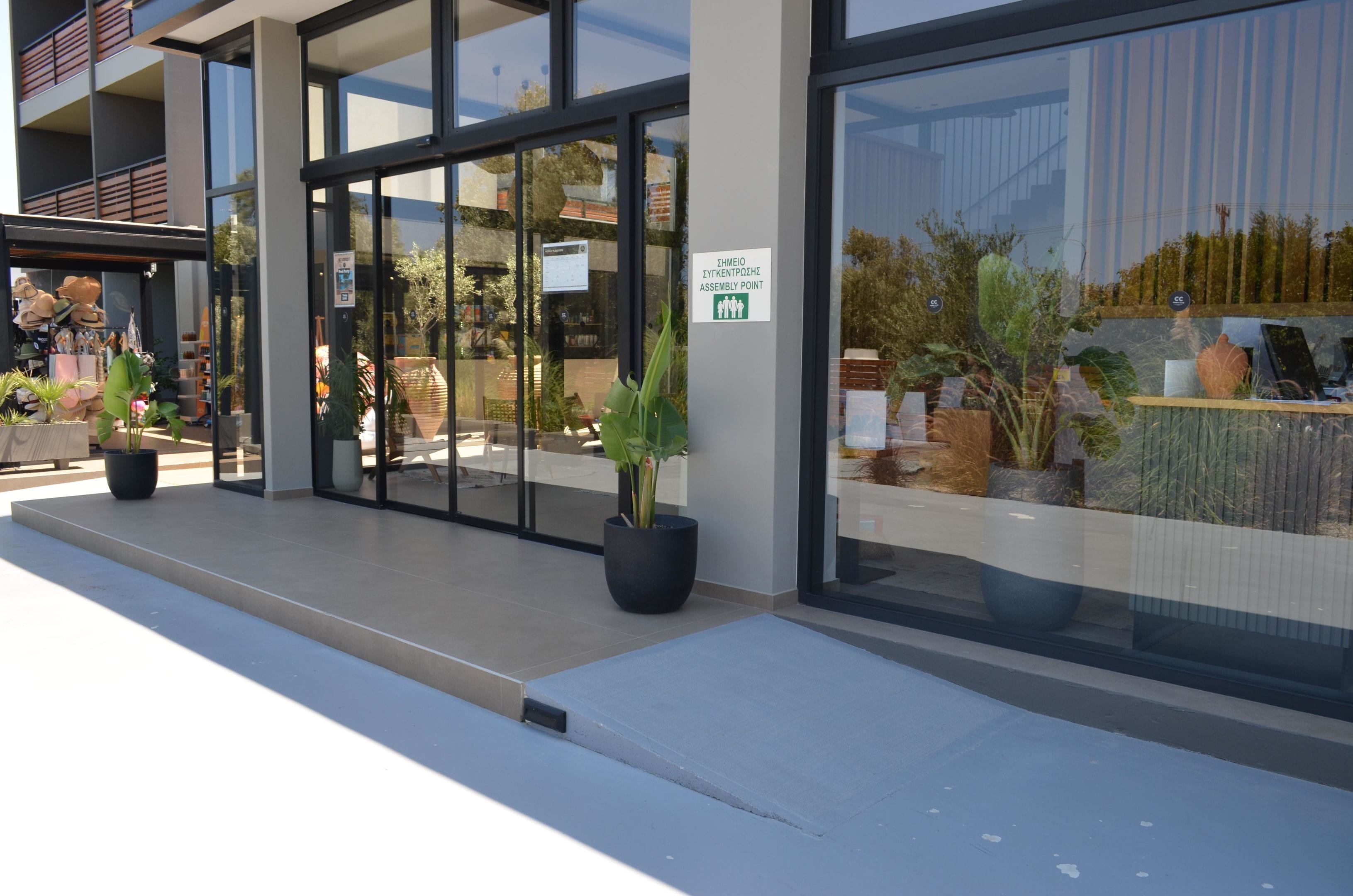 Un edificio moderno con grandes ventanales de vidrio, un cartel que indica punto de encuentro y plantas en macetas en el exterior.