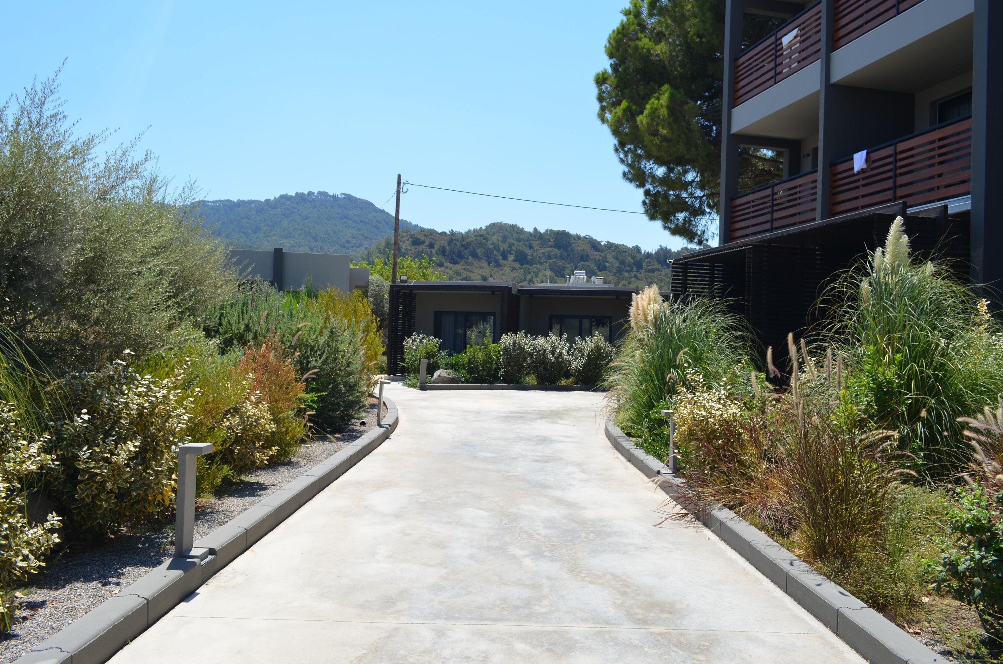 Un chemin mène à un bâtiment moderne doté de balcons, entouré de jardins aménagés et avec une montagne en arrière-plan.