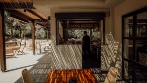 Ein Außenbereich mit Tischen und Stühlen, ein Barbereich mit Barkeeper und Blick auf Bäume.