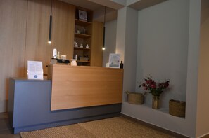 A reception desk with a wooden counter, a grey base, and a woven rug. There are shelves above the counter with various items and a framed picture. - Cook's Club Ialysos Rhodes (Rhodes)