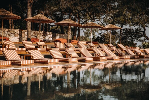 A poolside area with striped lounge chairs and thatched umbrellas, surrounded by stone walls and trees.