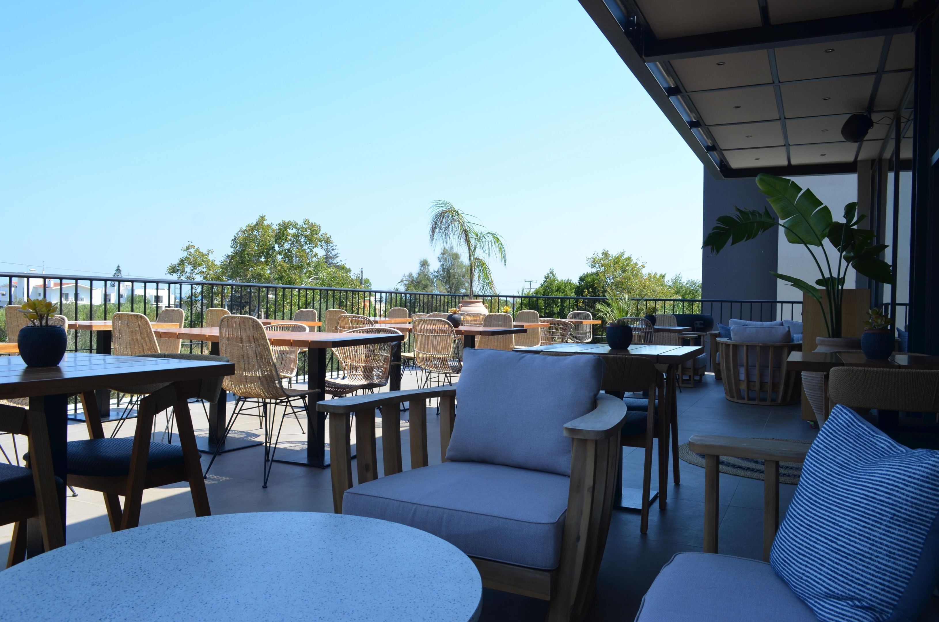 An outdoor patio area with round tables and wicker chairs, a covered seating area, and a view of palm trees and buildings in the background.