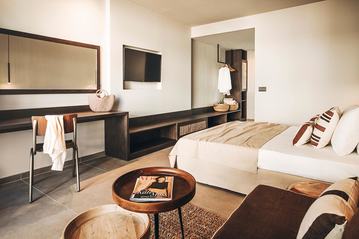 A modern hotel room with a large bed, a seating area with a magazine, a TV, and a large mirror.