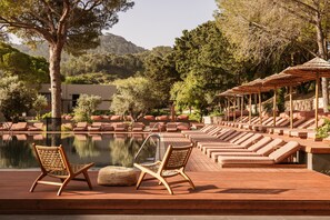 A poolside area with lounge chairs and umbrellas, surrounded by trees and a mountain backdrop.