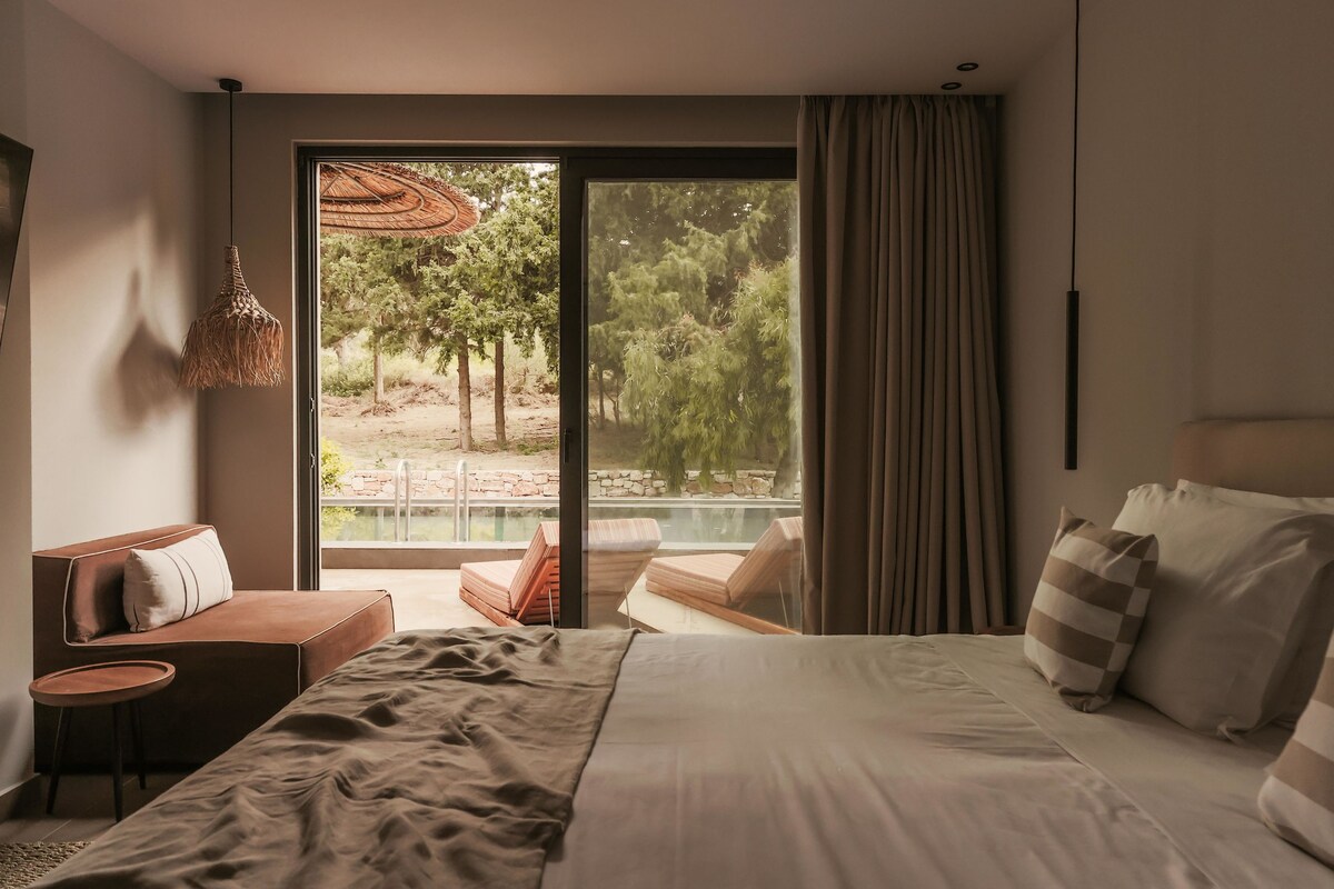 A modern hotel room with a large bed, a seating area with a sofa and chairs, a small table, and a view of a pool and outdoor area.