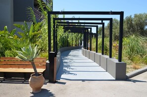 A pathway with a wooden bench and potted plants on the left, leading to a black metal structure with greenery.