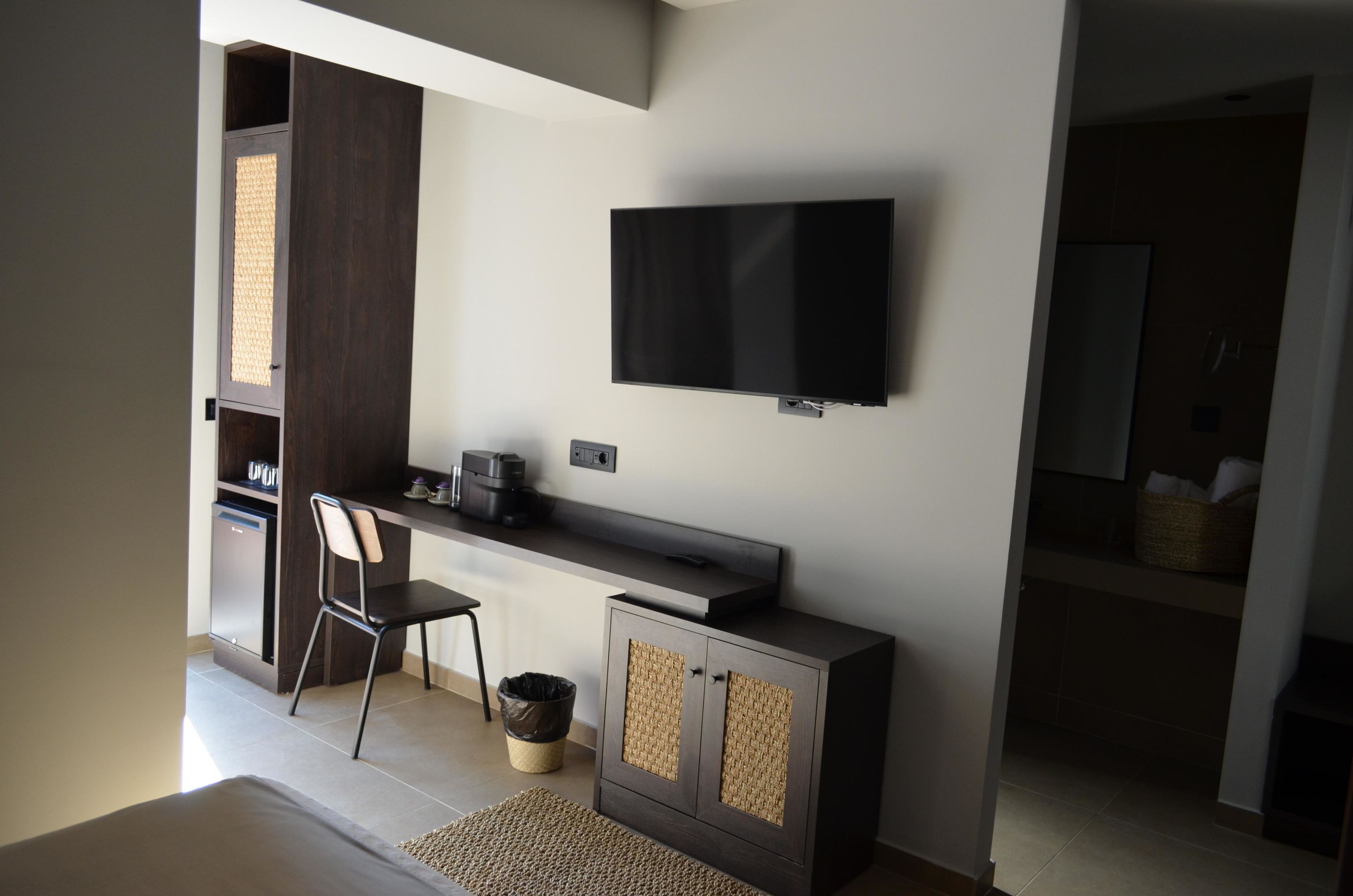 A modern room with a flat-screen TV mounted on the wall, a desk with a chair, a small cabinet, and a visible trash bin.