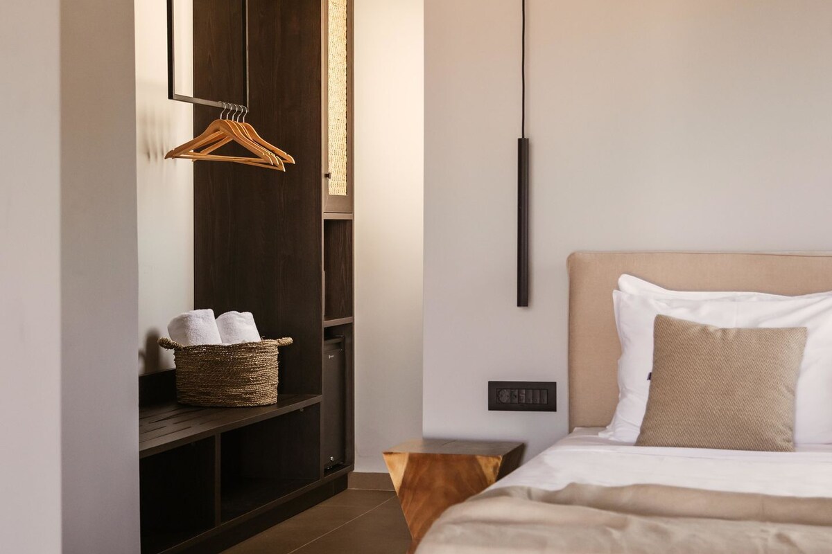 A modern hotel room with a large bed, a wooden bench with towels, a hanging light fixture, and a wardrobe.