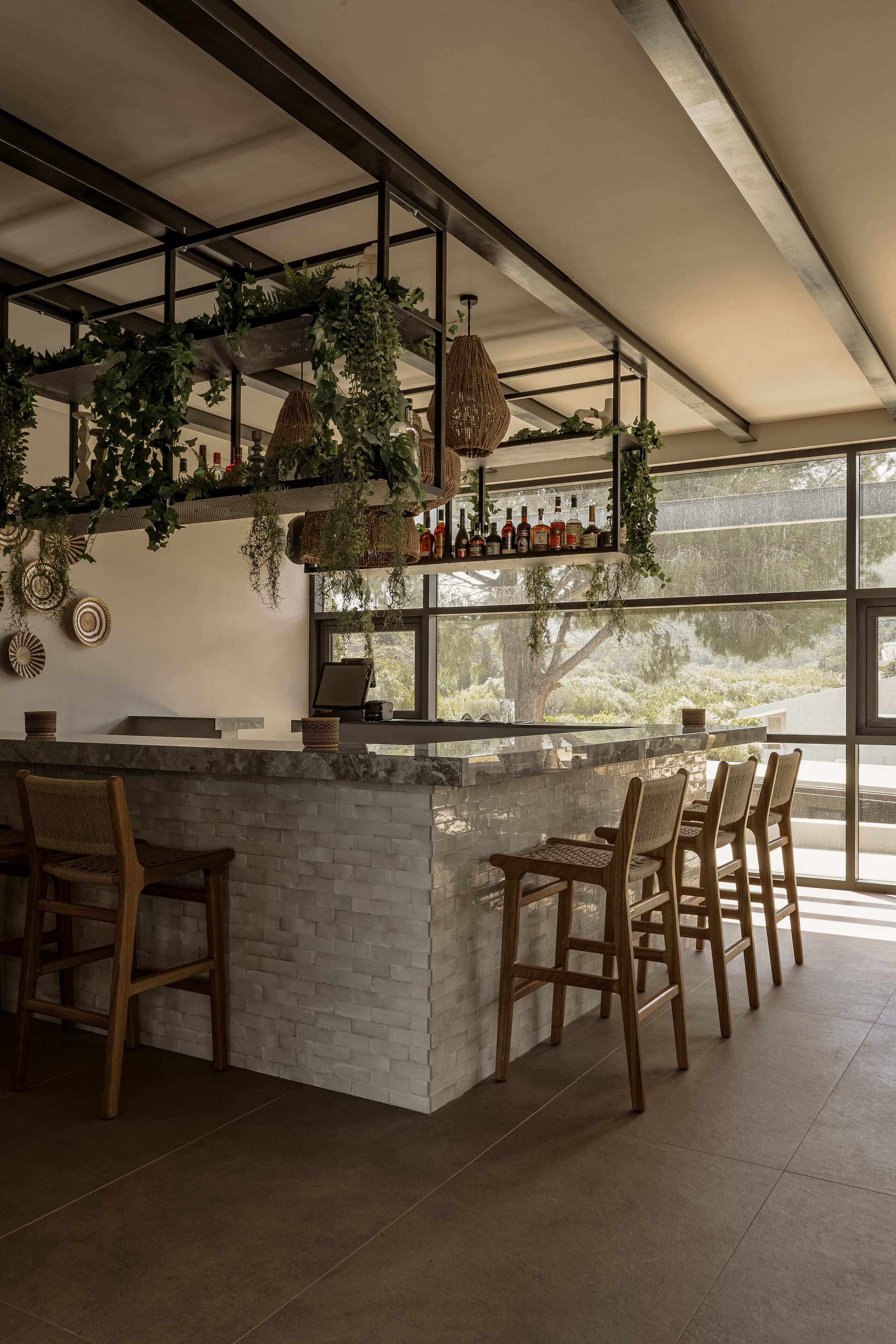 Una zona de bar con mostrador de piedra, sillas de madera, plantas colgantes y vistas a través de amplios ventanales.