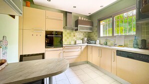 A kitchen with a dining table, cabinets, a stove, and a window with greenery outside. - Beautiful Villa 8 guests - Pool Access in Mandelieu (Mandelieu-la-Napoule)