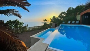 A swimming pool with a view of the sea at sunset, surrounded by a wooden fence and palm trees. - Sea-view villa for 11 guests with a heated infinity pool (Saint-Raphaël)