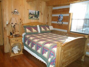 A wooden cabin bedroom with a quilted bedspread, a nightstand with a lamp, a small chest of drawers, and a window with blinds.