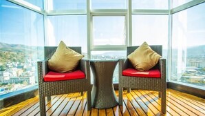 Twee rieten stoelen met rode kussens en een bijzettafeltje in een kamer met houten vloer en grote ramen met uitzicht op de stad.