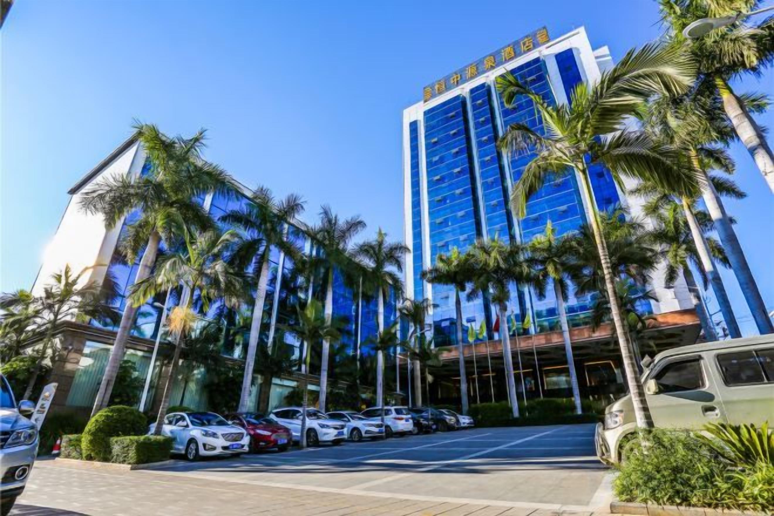 Un hotel con fachada de vidrio moderna, palmeras y un estacionamiento con varios automóviles.