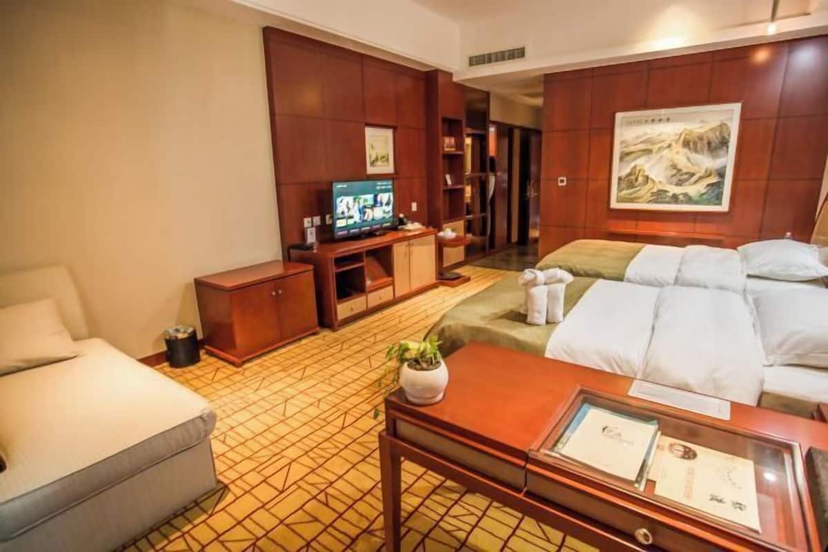 A hotel room with a bed, a TV, a desk, a chair, a small table with a plant, and a painting on the wall.
