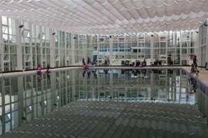 A large indoor swimming pool with a glass floor, surrounded by a modern architectural design with large windows and a patterned ceiling.