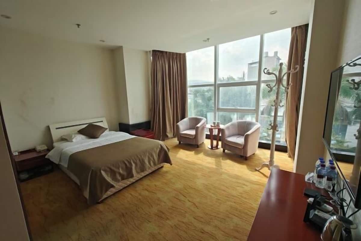 A hotel room with a bed, two armchairs, a small table, a TV, and a large window with curtains.