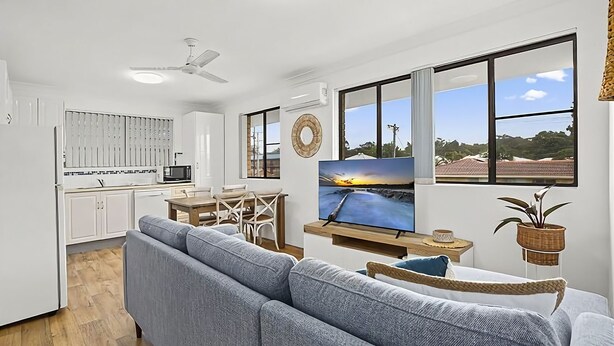 A modern kitchen with white cabinets, a dining area with a table and chairs, a large window with a view of the sea, and a living room with a sofa and a TV displaying a sunset scene.