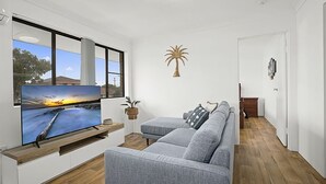 A modern living room with a flat-screen TV, grey sofa, wooden flooring, and a view of the outdoors through large windows.