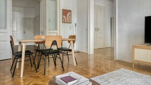 A modern living room with a wooden dining table, four chairs, a television on a wooden stand, and a bookshelf.