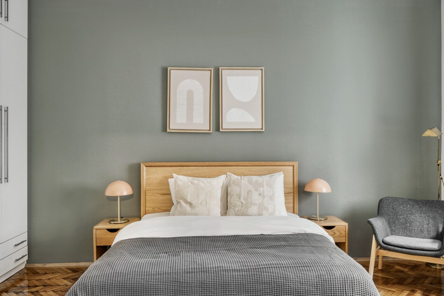 A modern bedroom with a wooden headboard, two bedside tables with lamps, a grey armchair, and two abstract wall art pieces.