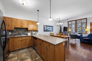 A modern kitchen with wooden cabinets, a central island, and a dining area with a blue sofa and a TV. - 2 bedroom condo with private hot tub, easy ski access (Sun Peaks)
