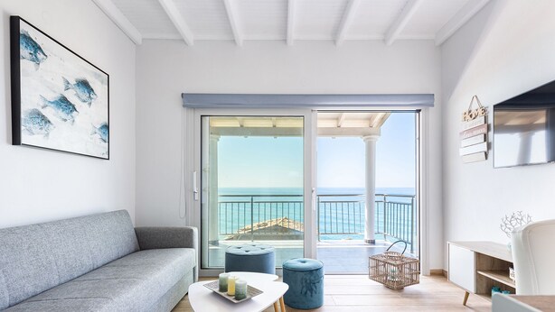 Un salon moderne avec un canapé gris, une table basse blanche ornée de bougies et une vue sur la mer par une porte vitrée.