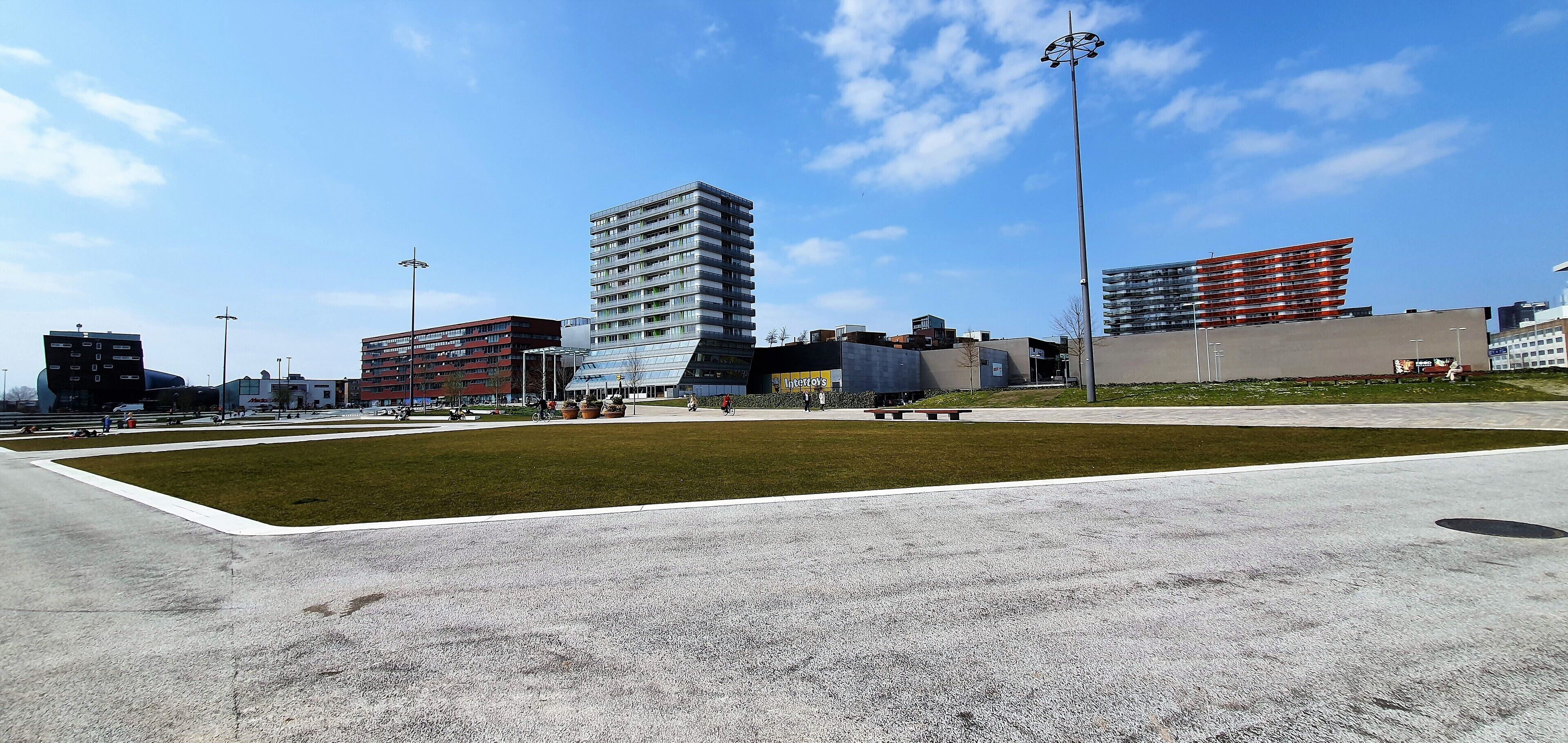 Une place urbaine moderne, dotée d’un espace central verdoyant, entourée d’immeubles contemporains aux styles architecturaux variés.