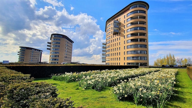 A modern apartment building with a curved design, a well-maintained garden with white flowers, and a clear blue sky. - Brand new apartment, next to Amsterdam and Floriade 2022, free parkingspace. (Almere)