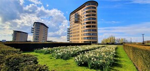 Een modern appartementencomplex met een golvend ontwerp, een verzorgde tuin met witte bloemen en een helderblauwe lucht.