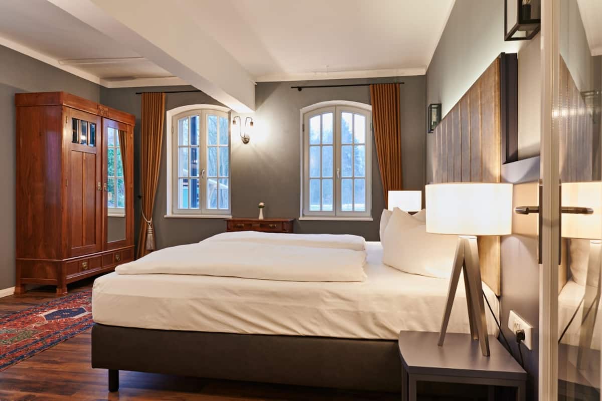 Ein Hotelzimmer mit einem großen Bett, einem Holzschrank, zwei Fenstern mit Vorhängen, einem Nachttisch mit Lampe und einem zusätzlichen Beistelltisch.