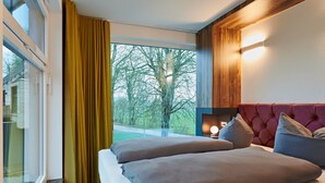 A hotel room with a large window, a bed with two pillows, a wooden headboard, and a nightstand with a lamp. - Bernsteinland Wendorf (Kuhlen-Wendorf)