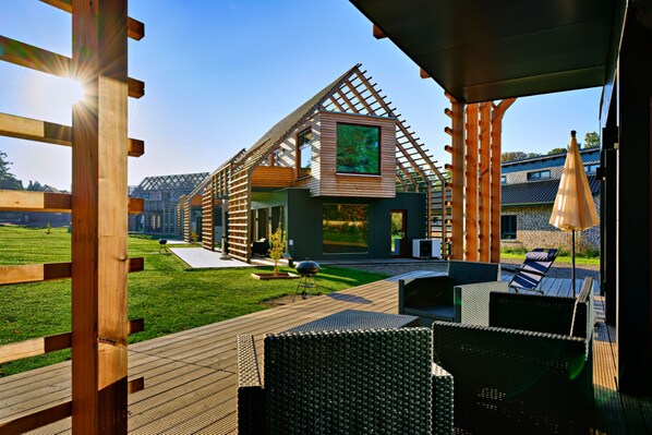 Una casa moderna con techo inclinado, terraza de madera, mobiliario exterior y vistas del entorno.
