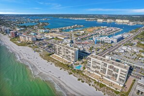 Vista aérea de una ciudad costera con un puerto deportivo, hoteles frente a la playa y un gran complejo hotelero con varias construcciones y zonas de estacionamiento.
