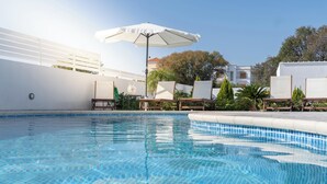 Una piscina con sillas de descanso y una sombrilla, rodeada de follaje y paredes blancas.