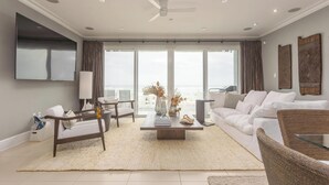 A modern living room with a flat-screen TV, white sofas, a coffee table, and a large window with curtains. - Contemporary Gulf Front Townhome Just Steps from the Water (Pensacola Beach)