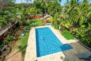 A rectangular swimming pool with clear blue water, surrounded by a stone deck. There are lounge chairs and a small garden area with various plants and a wooden fence in the background. - Private and Romantic Seaside Retreat - Just Twelve Steps to the Beach! (Hanalei)