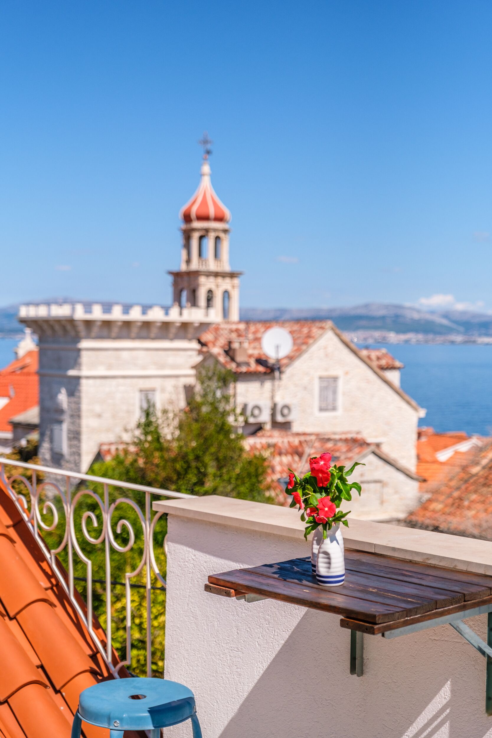 A balcony with a view of a historic building with a red dome and bell tower, a wooden table with a vase of flowers, and a blue stool.