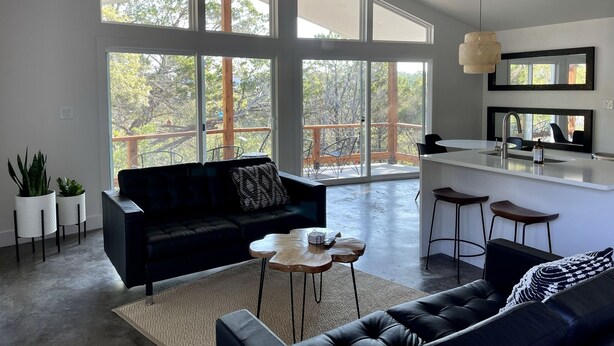 Amplia sala de estar con un sofá negro, una mesa de centro y un comedor con taburetes de bar. Amplias ventanas ofrecen vistas al exterior.