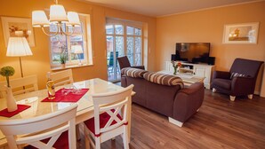 Un salon avec un canapé marron, une table à manger blanche avec des chaises, un téléviseur et une fenêtre donnant sur un paysage verdoyant.