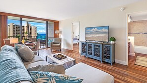 Amplia sala de estar con un ventanal que ofrece vistas del horizonte de la ciudad y del océano. Hay una zona de estar acogedora con un sofá, sillones, una mesa de centro y un televisor sobre un mueble.