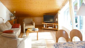 A cozy living room with a wooden ceiling, a sofa, a television, and a dining area with chairs. - Quietly situated apartment for max. 4 persons, balcony, garden, WLAN, washing machine (Prerow)
