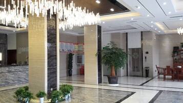 A hotel lobby with a reception desk, a large potted plant, and a modern chandelier.