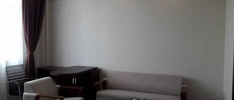 A modern room with a sofa, a small table, and a chair. There is a window with curtains, a radiator, and a trash bin.